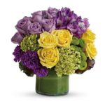 A round floral arrangement in a short green glass vase featuring lavender roses, bright yellow roses, deep purple carnations, green hydrangeas, and purple alstroemeria with lush green foliage.