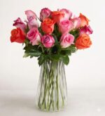 A vibrant bouquet of fresh roses in shades of pink, orange, and purple, arranged in a clear, vertically ribbed glass vase against a plain white background.