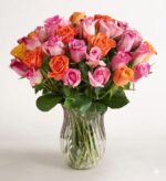 A large, vibrant bouquet of mixed pink, orange, and peach roses arranged in a clear, wavy glass vase against a white background.