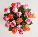 A top-down view of a vibrant rose bouquet featuring a mix of pink, orange, and lavender-tipped roses with green leaves against a light neutral background.