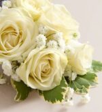 Close-up of an elegant wrist corsage featuring three ivory spray roses, clusters of white baby’s breath, and variegated green and cream leaves on a light background.