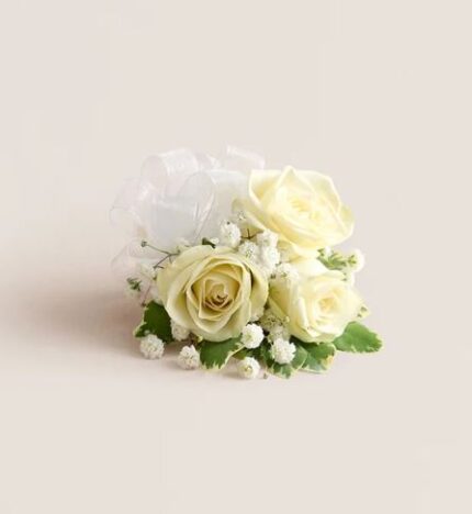 White spray rose wrist corsage with baby’s breath, variegated greenery, and a sheer white ribbon, on a soft cream background.