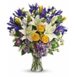 A large mixed spring bouquet in a clear glass vase featuring vibrant blue irises, yellow roses, white lilies, yellow tulips, purple hydrangea, small purple asters, and white filler flowers with green foliage.