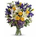 Vibrant premium bouquet in a clear glass vase featuring white lilies, yellow roses, deep purple irises, purple asters, lavender hydrangeas, and white filler flowers with green accents.