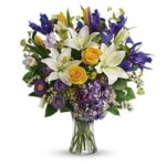 A tall glass vase filled with a vibrant mixed bouquet of white lilies, yellow roses, blue irises, purple asters, lavender hydrangea, and light purple lisianthus with lush green foliage.