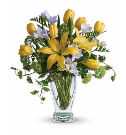 Clear glass vase with ivy accents holding a bright spring bouquet of yellow tulips, large yellow Asiatic lilies, white freesias, and green hydrangea blooms, arranged against a white background.