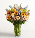A lush and colorful mixed flower bouquet in a clear glass vase, featuring peach roses, yellow sunflowers, blue delphiniums, and pink alstroemeria against a white background.