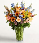 A tall, lush floral arrangement in a clear glass vase featuring peach roses, yellow sunflowers, blue delphiniums, and pink alstroemeria against a plain white background.