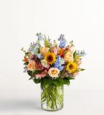 A vibrant "Spring Sensation" floral arrangement in a clear cylindrical glass vase featuring yellow sunflowers, peach roses, light blue delphiniums, and pink alstroemeria against a white background.