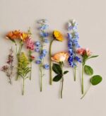 A flat-lay arrangement of various colorful spring flowers, including blue delphiniums, orange alstroemerias, a yellow sunflower, and a peach rose, laid out vertically against a neutral beige background.