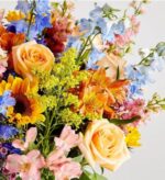 A vibrant and dense spring flower bouquet featuring peach roses, a bright yellow sunflower, light blue delphiniums, orange alstroemeria, and pink snapdragons with yellow solidago accents.