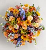 A vibrant and dense top-down view of a spring flower bouquet featuring peach roses, bright yellow sunflowers, blue delphiniums, and orange alstroemeria with green foliage.