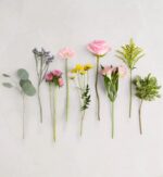 Eight assorted flower and greenery stems laid flat on a white marble background. From left to right: eucalyptus, purple statice, pink chrysanthemums, light pink gerbera daisy, yellow billy buttons, pale pink rose, pink alstroemeria with greenery, yellow solidago, and variegated pittosporum.