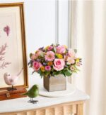 A pastel spring flower arrangement in a square wooden box, featuring pink roses, light pink gerbera daisies, and small yellow and purple accent blooms with eucalyptus greenery. The box sits on a white marble table beside two small ceramic bird figurines and a gold-framed botanical print. Soft natural light filters through a sheer curtain in the background.