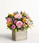 A square wooden box vase filled with a spring bouquet of light pink roses, pink gerbera daisies, yellow button mums, lavender sprigs, pink carnations, and green foliage on a white background.