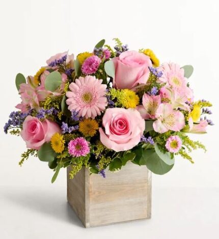 A square light wood box filled with a vibrant spring flower arrangement featuring pink roses, light pink gerbera daisies, magenta button mums, yellow craspedia, pink alstroemeria, purple statice, and sprigs of eucalyptus greenery.
