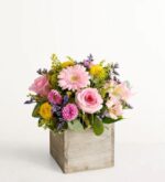 Square rustic wood planter box filled with a spring flower arrangement of pink roses, light pink gerbera daisy, pink chrysanthemums, yellow button mums, purple statice, and fresh greenery against a white background.