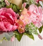 Close-up of a floral centerpiece in a white ribbed ceramic vase, featuring large hot pink and blush pink peonies, peach stock flowers, small pink filler blooms, and soft green lamb’s ear leaves, with a cream pillar candle in the background.
