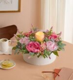 A low, round white ribbed ceramic vase on a peach-colored table holds a lush floral centerpiece with large pink peonies, blush roses, light purple stock flowers, and green eucalyptus leaves. A glowing flameless candle sits in a glass holder at the center. A white mug, a small plate with lemon slices, and a pair of eyeglasses are nearby on the table.