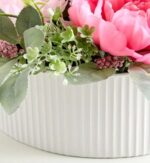 Close-up of a white ribbed oval vase filled with large pink peony blooms, lamb's ear greenery, small green blossoms, and clusters of mauve accent flowers.