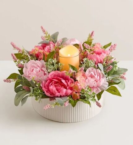 Round white ribbed ceramic bowl filled with pink peonies, pink ranunculus, small pink blossoms, and green foliage, arranged around a glowing LED candle in a glass holder.