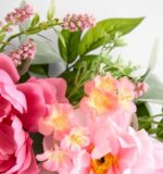 Close-up of an artificial spring wreath featuring large soft pink peonies, pale peach-pink blossoms with yellow centers, small clusters of tiny pink filler flowers, and lush green leaves on a white background.