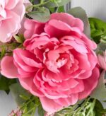Close-up of a vibrant pink artificial peony flower with layered petals, surrounded by soft pink buds, small pink filler flowers, and green eucalyptus-style leaves on a spring wreath