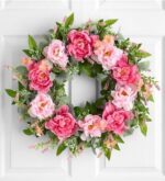Round door wreath of large pink and blush peonies with small peach blossoms and green eucalyptus leaves, hanging on a white paneled door.