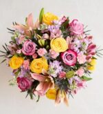 A vibrant, top-down view of a dense floral bouquet featuring yellow roses, pinkish-purple roses, light pink lilies, purple stock flowers, and blue thistles. Title: Springtime Blossoms for Mom