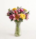 A vibrant and diverse bouquet of spring flowers featuring pink lilies, yellow roses, purple roses, and blue thistles in a clear fluted glass vase.