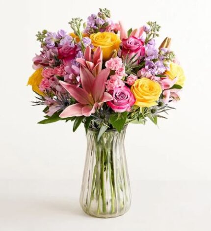 A vibrant and lush floral bouquet in a clear glass vase featuring pink lilies, yellow and pink roses, lavender stocks, and small pink carnations against a white background.