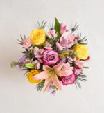 A top-down view of a vibrant spring bouquet featuring yellow roses, pink lilies, purple carnations, and blue thistles against a neutral light background.