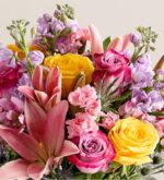 A vibrant and dense bouquet of spring flowers featuring pink lilies, yellow roses, purple-and-pink bicolored roses, pink carnations, lavender stock flowers, and blue sea holly accents.