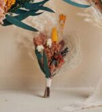 Small terracotta-toned dried flower boutonnière with white flowers, rust and mustard accents, pampas grass, and eucalyptus leaves, wrapped in ivory ribbon