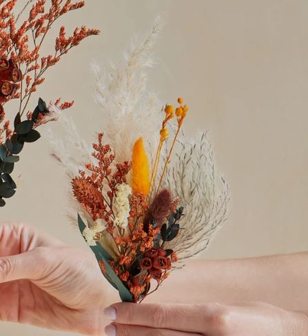 Close-up of hands holding a terracotta-toned dried flower boutonnière featuring pampas grass, bunny tails, preserved roses, eucalyptus leaves, and small orange and cream accent blooms against a soft beige background.