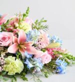 A lush floral centerpiece with soft pink lilies, roses, light blue delphiniums, and pale green carnations set against a neutral background.