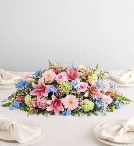 A lush, horizontal floral centerpiece featuring pink lilies, roses, and gerbera daisies accented with light blue delphiniums and green carnations on a white table.
