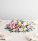 A lush, horizontal floral centerpiece on a white dining table, featuring pink lilies, pink gerbera daisies, light green carnations, and blue delphiniums surrounded by four place settings with white napkins.