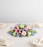 A horizontal floral centerpiece featuring pink lilies, gerbera daisies, roses, light green carnations, and blue delphiniums arranged on a white tablecloth with four place settings.
