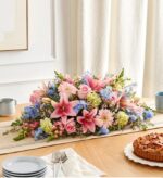 A lush, horizontal floral centerpiece featuring a mix of pink lilies, soft pink roses, gerbera daisies, and light blue delphiniums arranged on a wooden dining table with a neutral table runner.