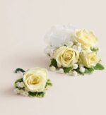 Ivory spray rose wrist corsage with white ribbon and baby’s breath next to matching single rose boutonniere, both accented with variegated greenery, displayed on a light beige background.