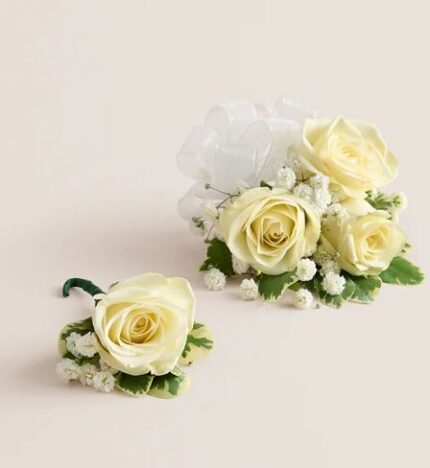 Ivory spray rose wrist corsage with white ribbon and baby’s breath next to matching single rose boutonniere, both accented with variegated greenery, displayed on a light beige background.