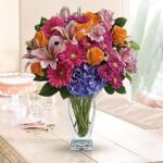 Vibrant mixed flower bouquet in a clear glass vase on a light wood table. Features hot pink gerbera daisies, orange roses, lavender lilies, purple hydrangea, and light pink alstroemeria, with green stems visible through the water.