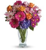 A vibrant floral bouquet in a clear glass vase featuring pink gerbera daisies, orange roses, purple hydrangea, light pink lilies, lavender alstroemeria, and hot pink chrysanthemums, arranged with lush greenery.