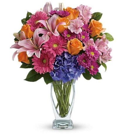 A vibrant mixed flower bouquet in a clear glass vase, featuring pink gerbera daisies, purple hydrangea, orange roses, pink lilies, light pink alstroemeria, and small magenta chrysanthemums.