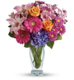 A vibrant mixed flower bouquet in a clear glass vase, featuring hot pink gerbera daisies, orange roses, light pink lilies, purple hydrangea, and purple daisies with yellow centers.