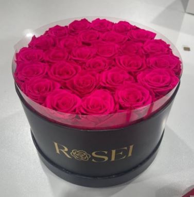 A circular matte black luxury flower box filled with vibrant hot pink preserved roses, featuring the gold logo "ROSEI" on the front.
